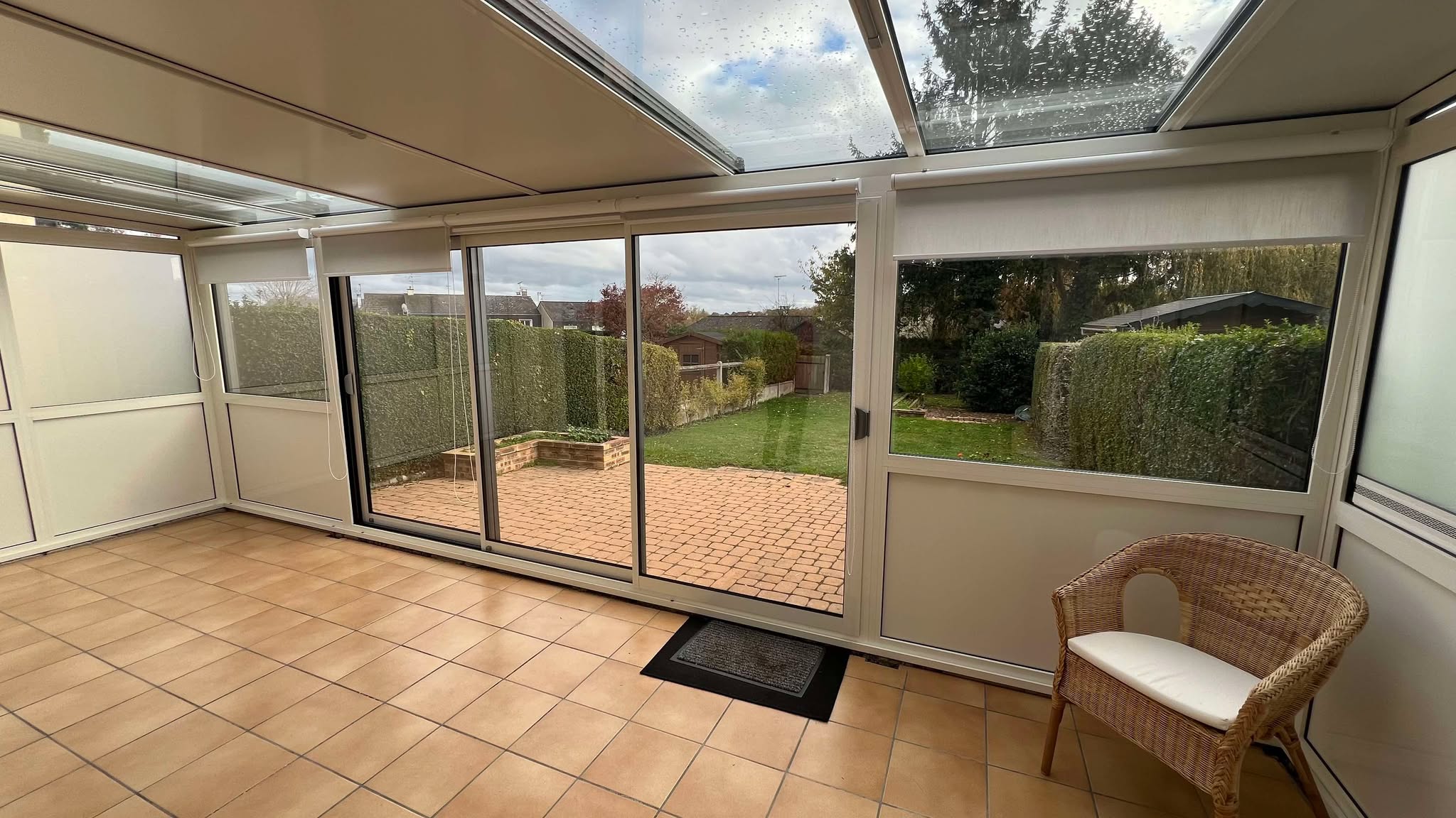 Véranda en PVC avec baies vitrées et stores, sol en carrelage.