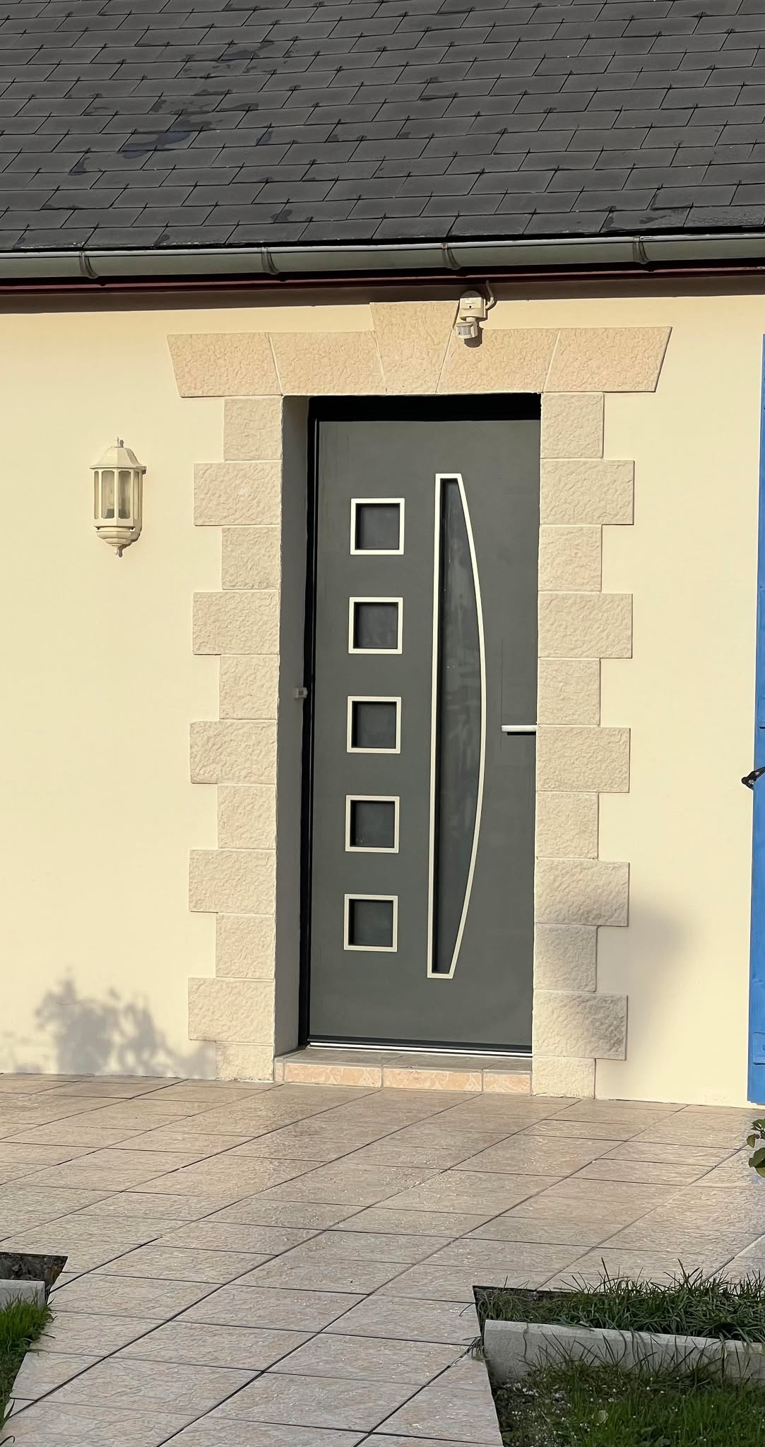 Porte d'entrée en métal gris avec éléments décoratifs en verre, encadrée de pierre.