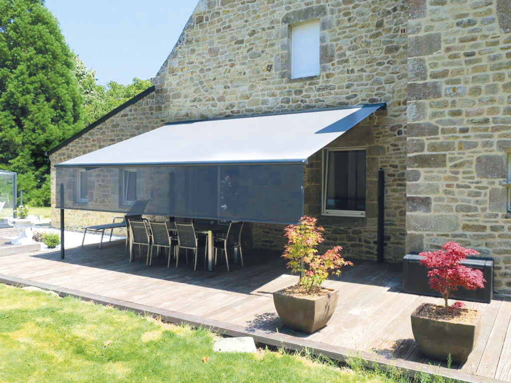 Store en toile grise installé au-dessus d'une terrasse, fixé à la facade de la maison.