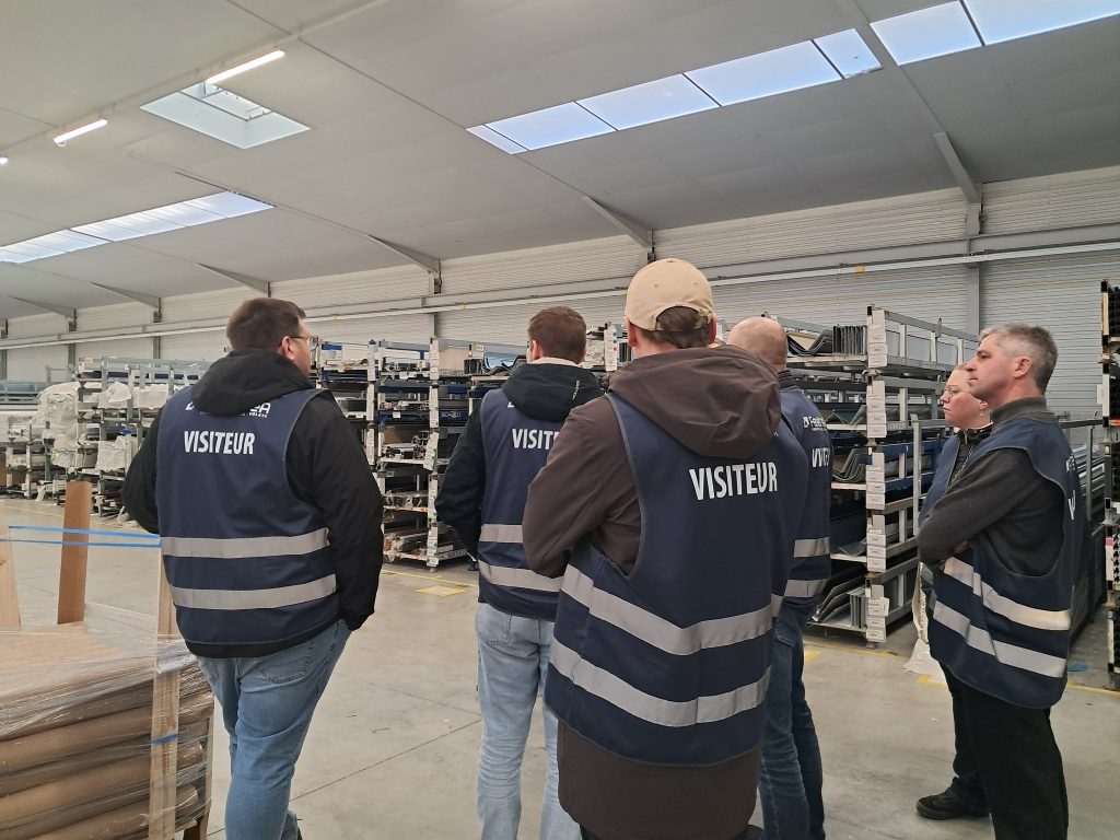 Visiteurs portant des gilets, observant des étagères avec des menuiseries et divers matériaux de construction.